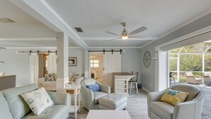 Uma sala de estar espaçosa com ventoinha no teto, uma área de jantar com mesa e cadeiras, e vista através de amplas janelas.