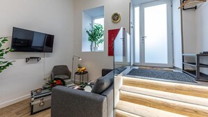 Una sala moderna con un sofá gris, un televisor de pantalla plana, una mesita con fruta, una planta en maceta y una escalera que sube a un nivel superior.