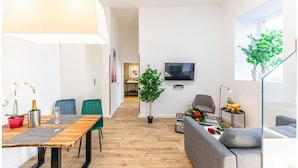 Una sala moderna con una mesa de comedor de madera, un sofá gris, un televisor montado en la pared y un ventanal con una planta.