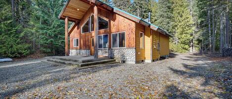 Une cabane en bois, avec des fondations en pierre, de grandes fenêtres et une allée en gravier, le tout entouré d’arbres.