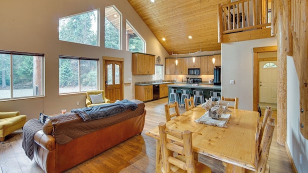 A spacious living area with a large wooden dining table, a sofa, and a kitchen in the background. - 3 Mi to Lake Easton Sno-park: Family Cabin Escape! (Easton)