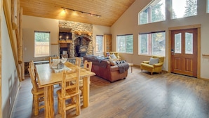 A spacious living area with a wooden dining table, a leather sofa, and a yellow armchair. A stone fireplace and a large window are visible. - 3 Mi to Lake Easton Sno-park: Family Cabin Escape! (Easton)