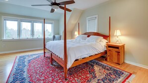Ein Bett mit Baldachin, dessen Holzrahmen mit weißer Bettwäsche bezogen ist, ein Deckenventilator, ein Nachttisch mit Lampe und ein Teppich mit blau-rotem Muster.