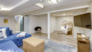 A modern living room with a blue sofa, a wicker basket, a large flat-screen TV, and a staircase leading to an upper level. - Charming Arnold Home w/ Patio & Chicken Coop! (Arnold)