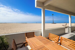 Una mesa y sillas de madera en un balcón con vista a una playa, una palmera y una valla.