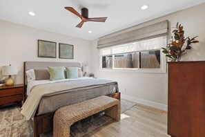 A bedroom with a bed, bedside tables, a ceiling fan, a window with blinds, and a decorative plant.