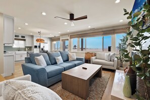 Una sala de estar moderna con un sofá azul, una mesa de centro de madera y vista a la playa a través de amplias ventanas.