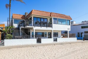 A two-story beachfront property with a balcony, a stone chimney, and a clear blue sky.
