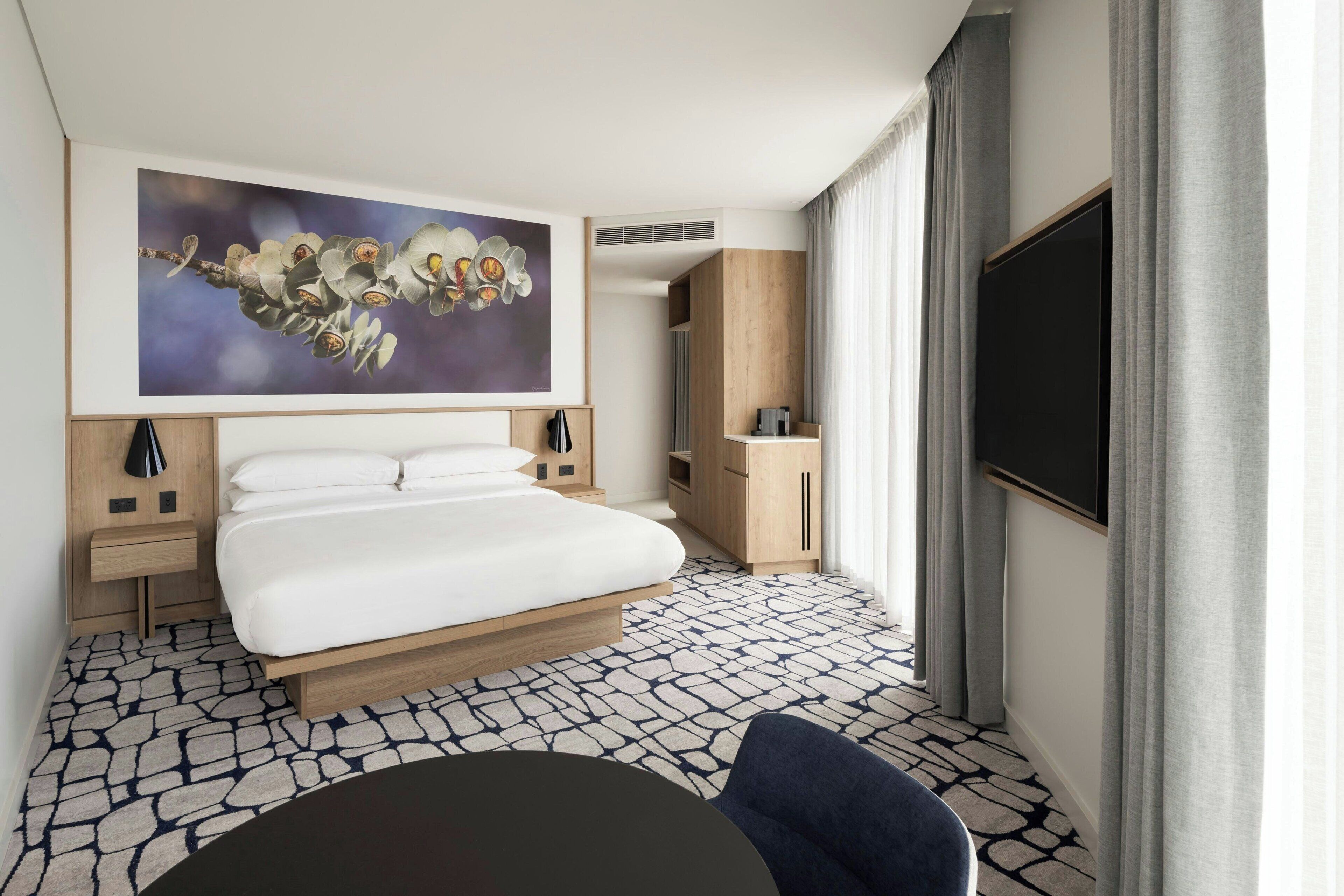 Una habitación de hotel moderna con una cama grande, un televisor de pantalla plana, un escritorio y un amplio ventanal con cortinas.