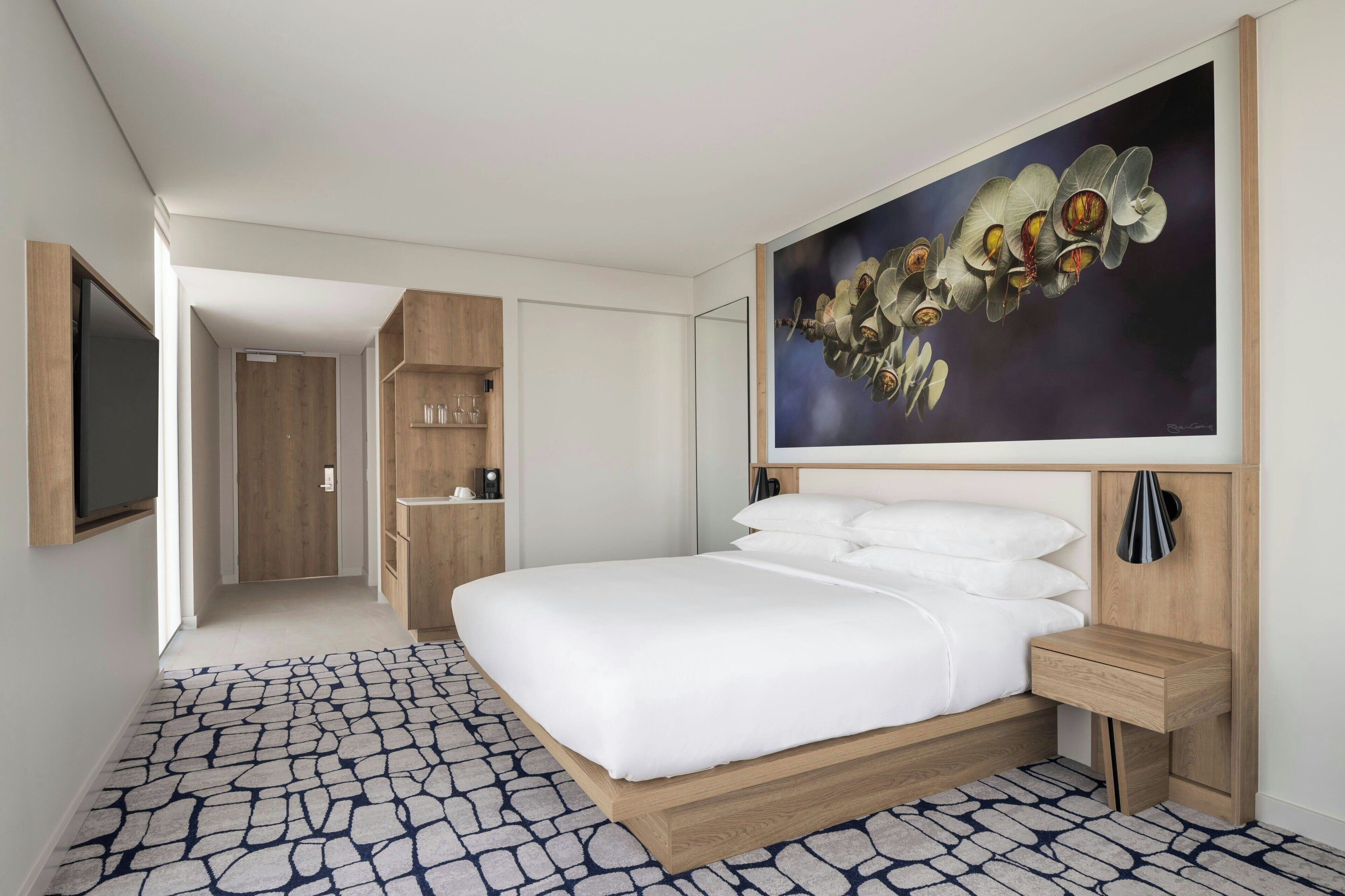 Habitación de hotel con una cama grande, cabecera de madera, mesita de noche y televisor montado en la pared.