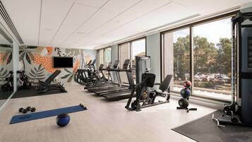 Un gimnasio con cintas de correr, máquinas de pesas y pelotas de ejercicio, ventanas amplias con vistas a árboles y coches estacionados.