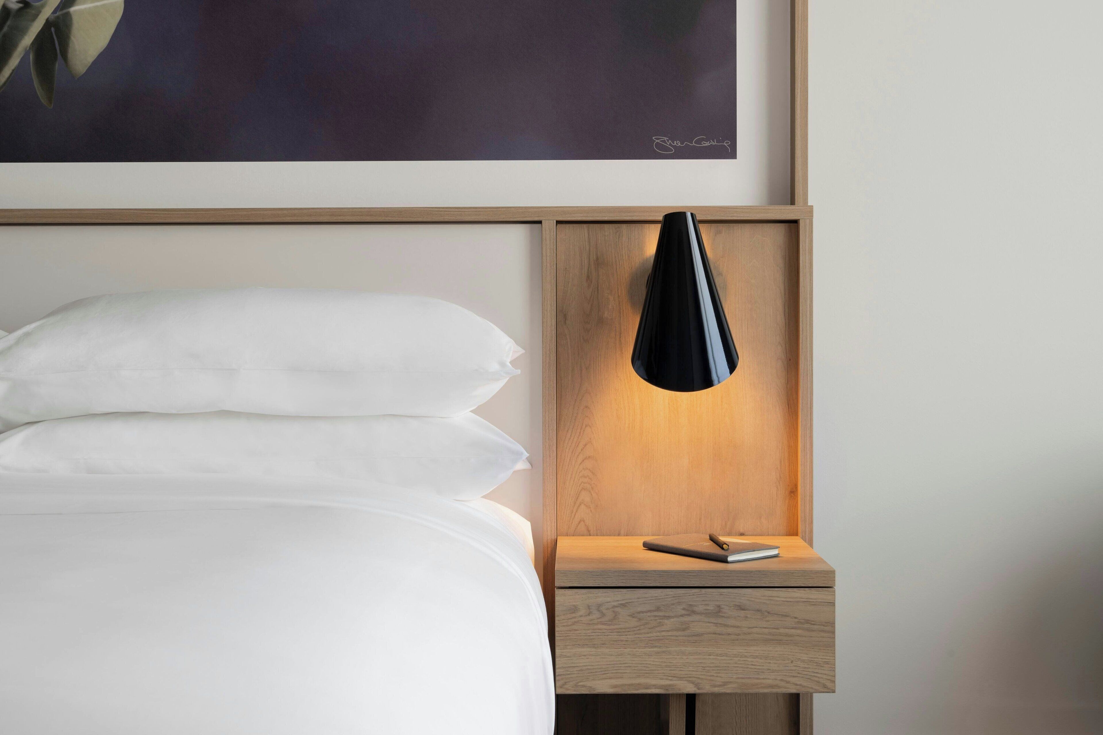 Ein ordentlich bezogenes Bett mit weißen Leinen und Kissen, ein Nachttisch mit einer schwarzen Lampe und ein gerahmtes Kunstwerk über dem Bett.