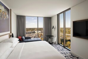 A modern hotel room with a large bed, a small table, a chair, and a TV. The room has large windows offering a city view.