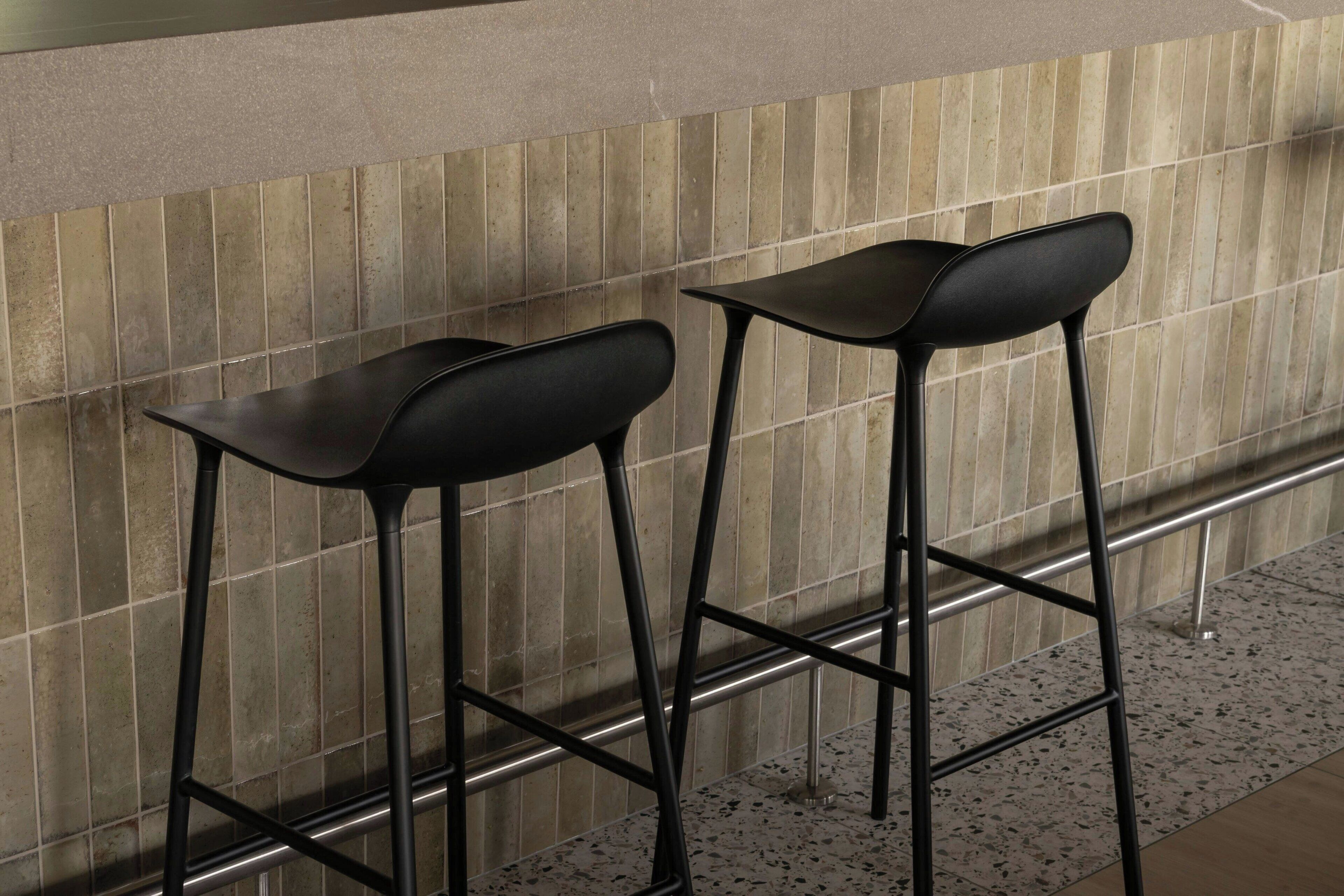 Dos taburetes negros con estructura metálica y una pared de azulejos al fondo.