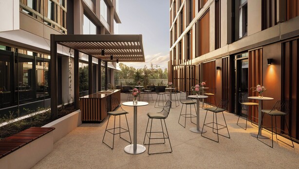 Un patio al aire libre con varias mesas y sillas, rodeado de edificios modernos con grandes ventanales y una pérgola que da sombra.