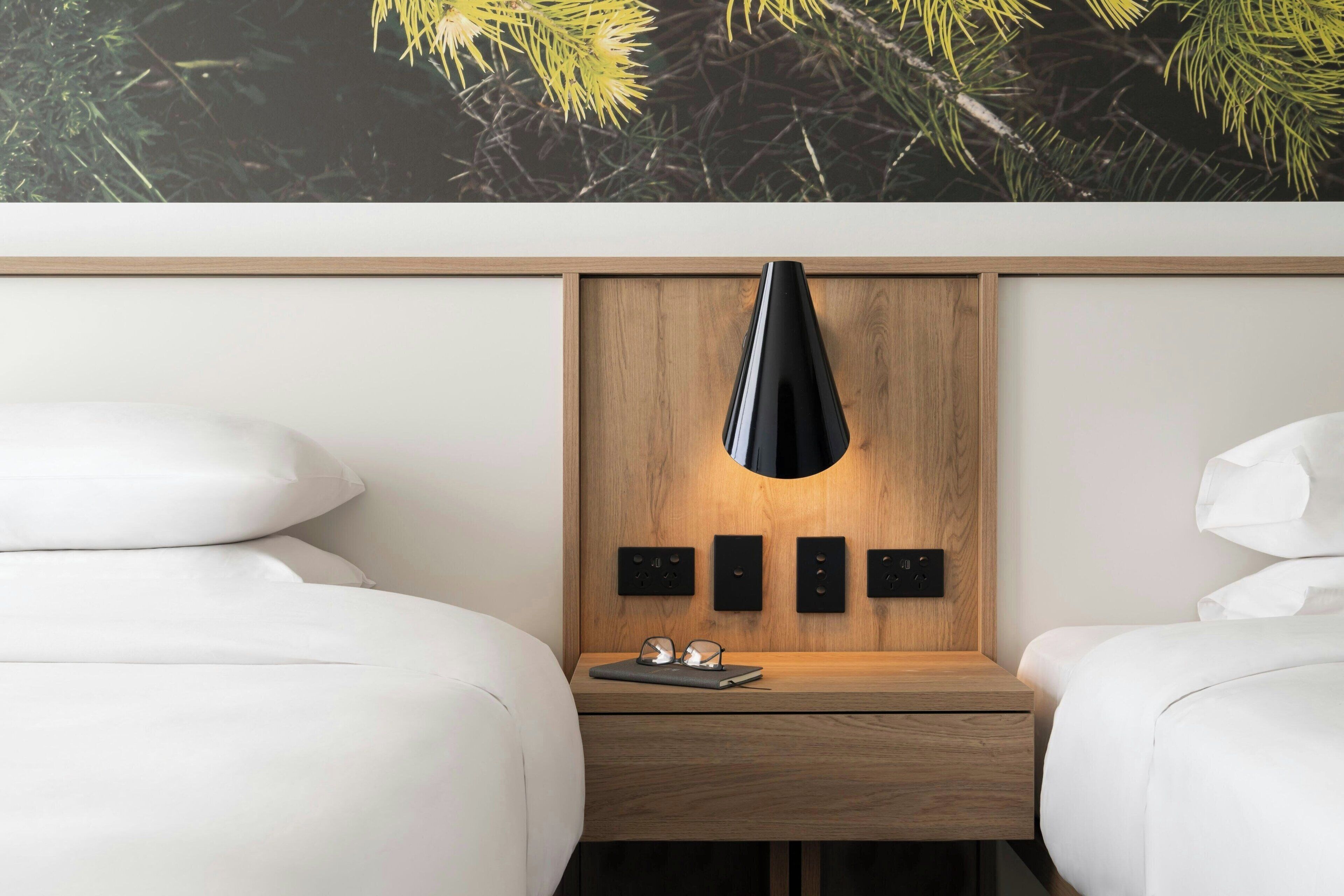 A modern hotel room with two beds, a wooden nightstand, a black pendant lamp, and a wall-mounted control panel.