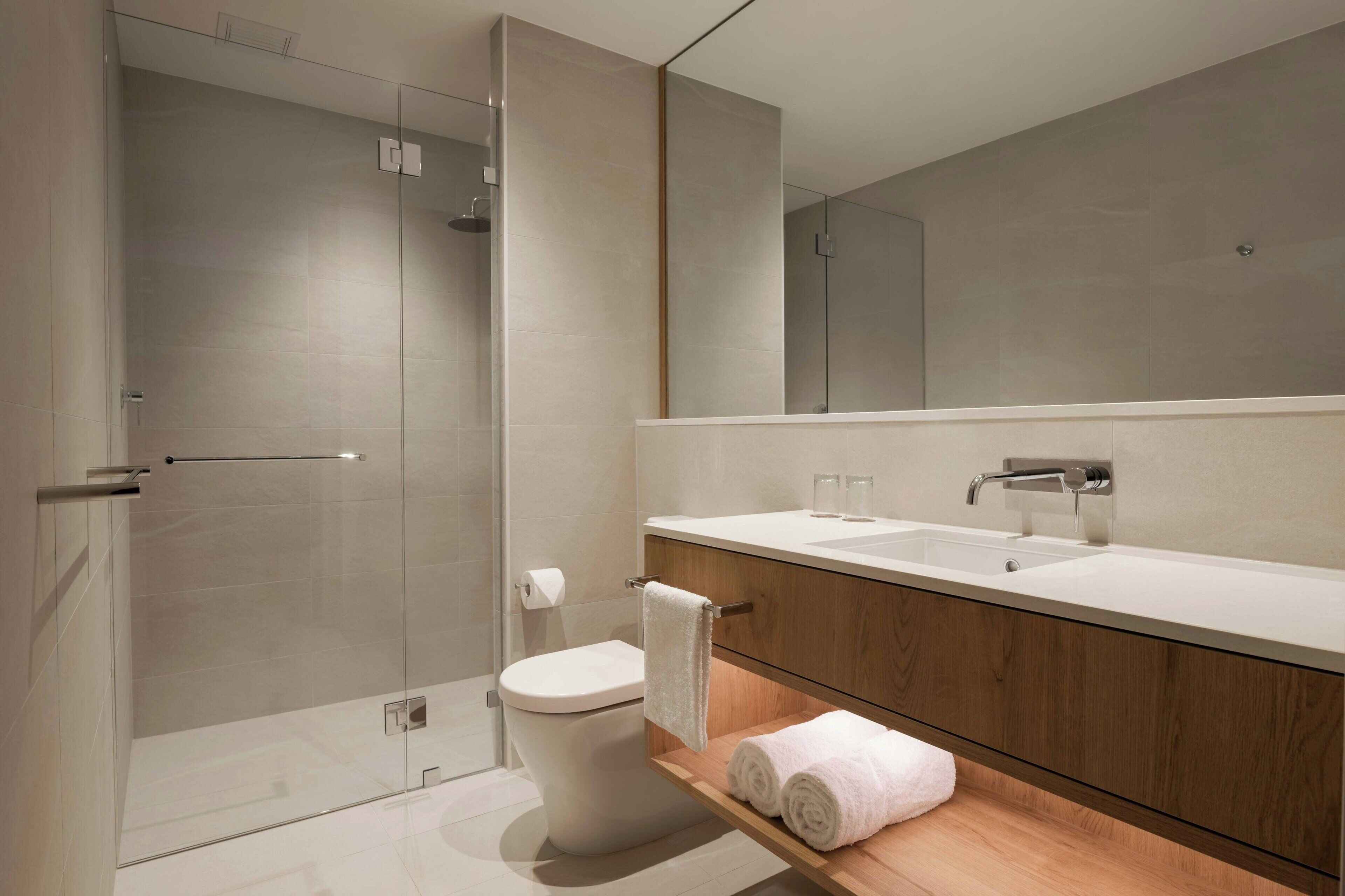 Une salle de bain moderne avec une douche à parois de verre, des toilettes blanches, un meuble-lavabo en bois et un grand miroir.