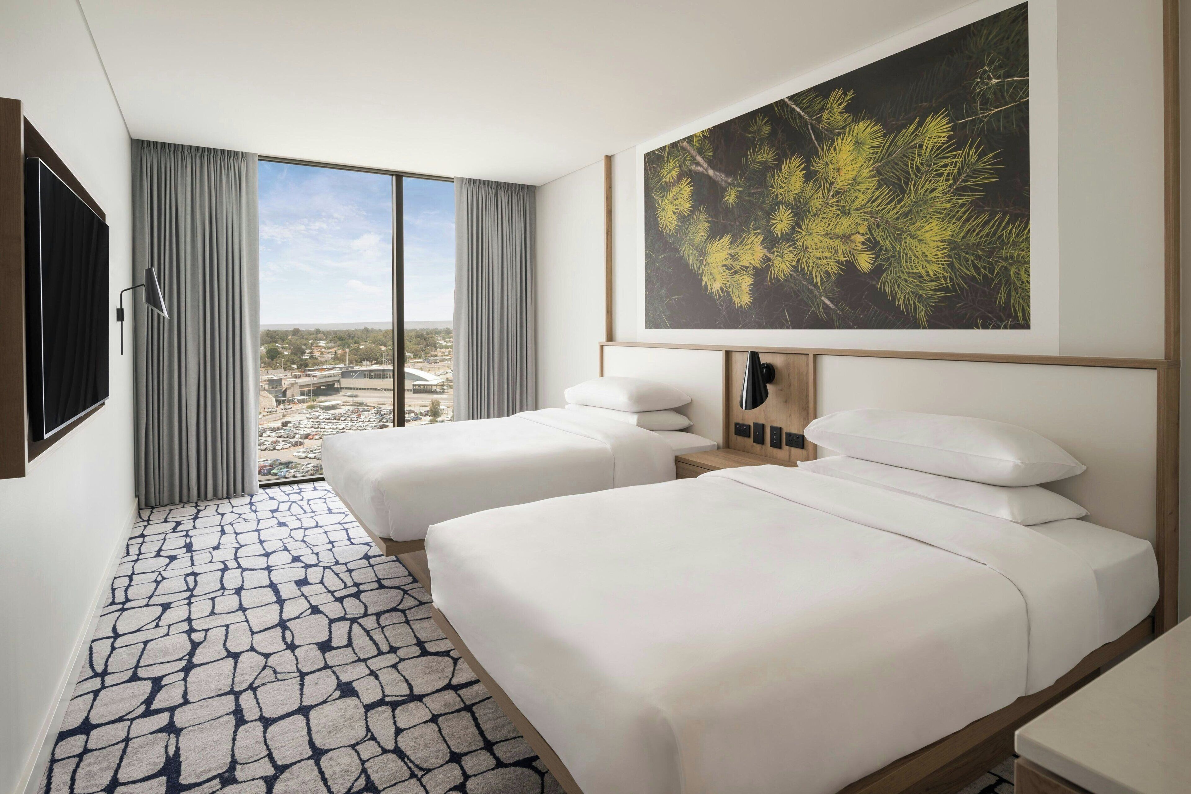 Une chambre d’hôtel avec deux lits, une grande fenêtre avec des rideaux, une vue sur la ville et une œuvre d’art encadrée au mur.