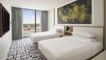 A hotel room with two beds, a large window with curtains, a city view, and a framed artwork on the wall.