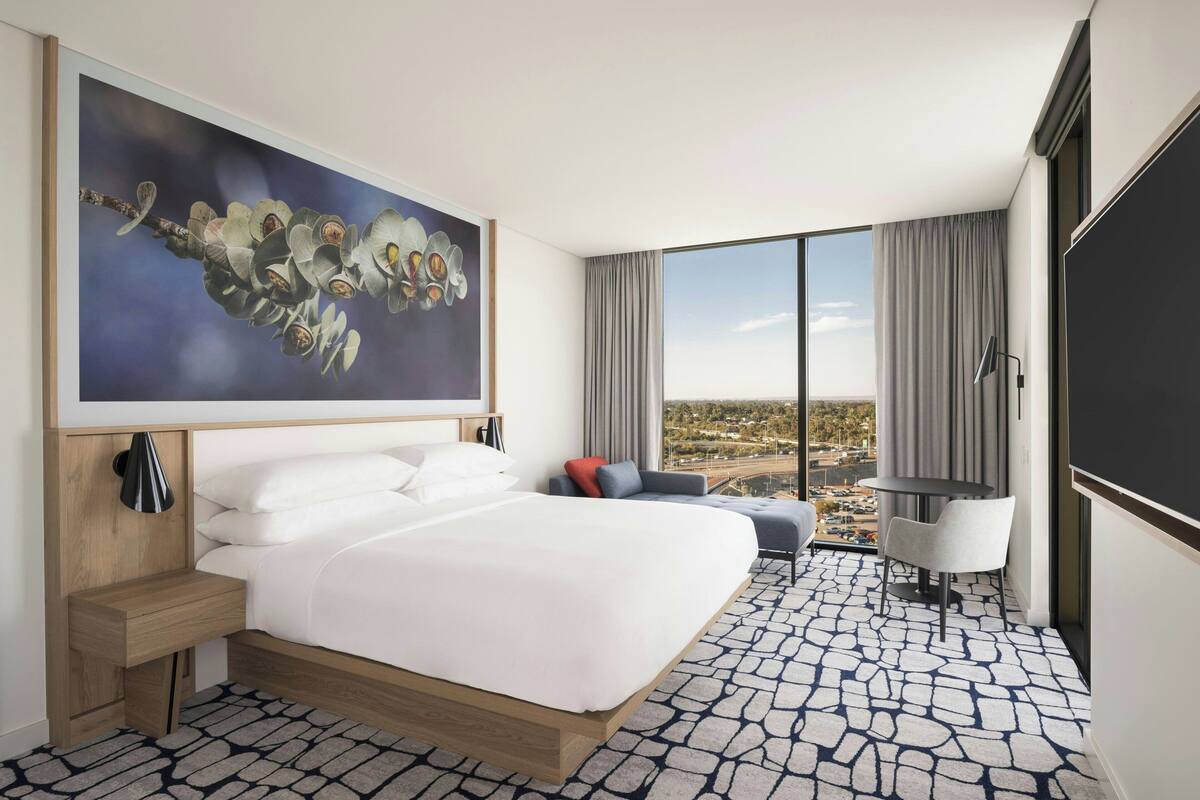 Quarto de hotel moderno com uma cama grande, uma escrivaninha, uma cadeira e uma janela ampla com vista para a cidade.