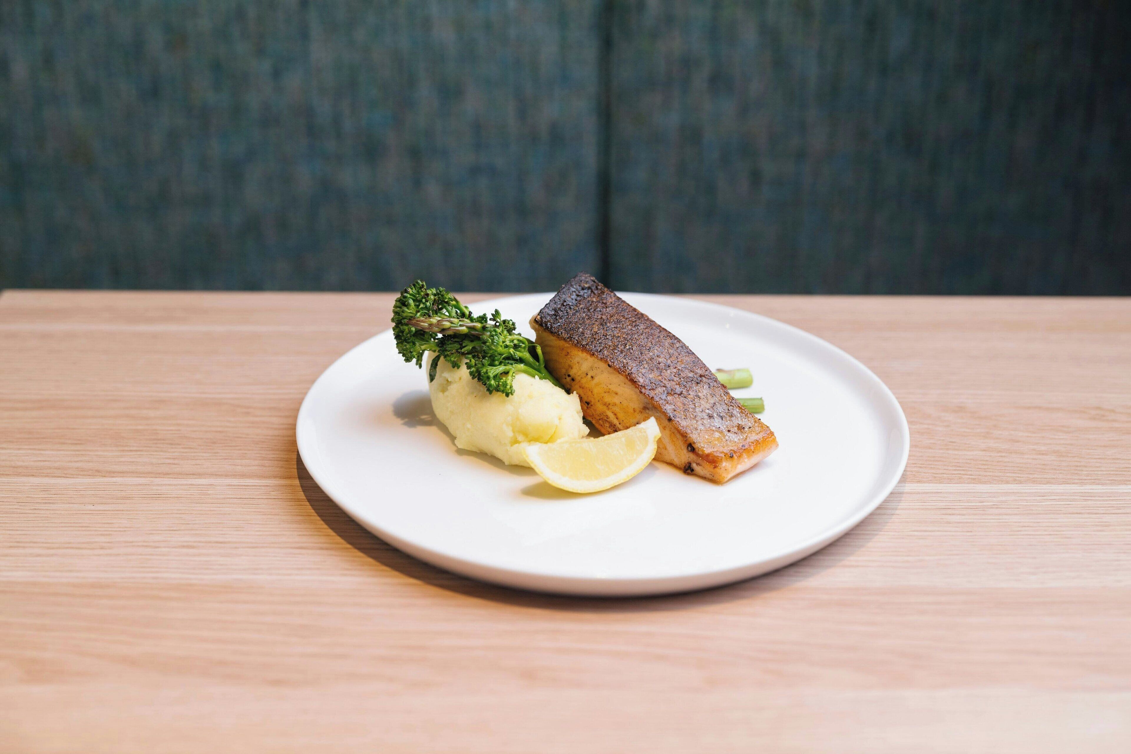 A plate with a piece of fish, mashed potatoes, and a lemon wedge on a wooden table.
