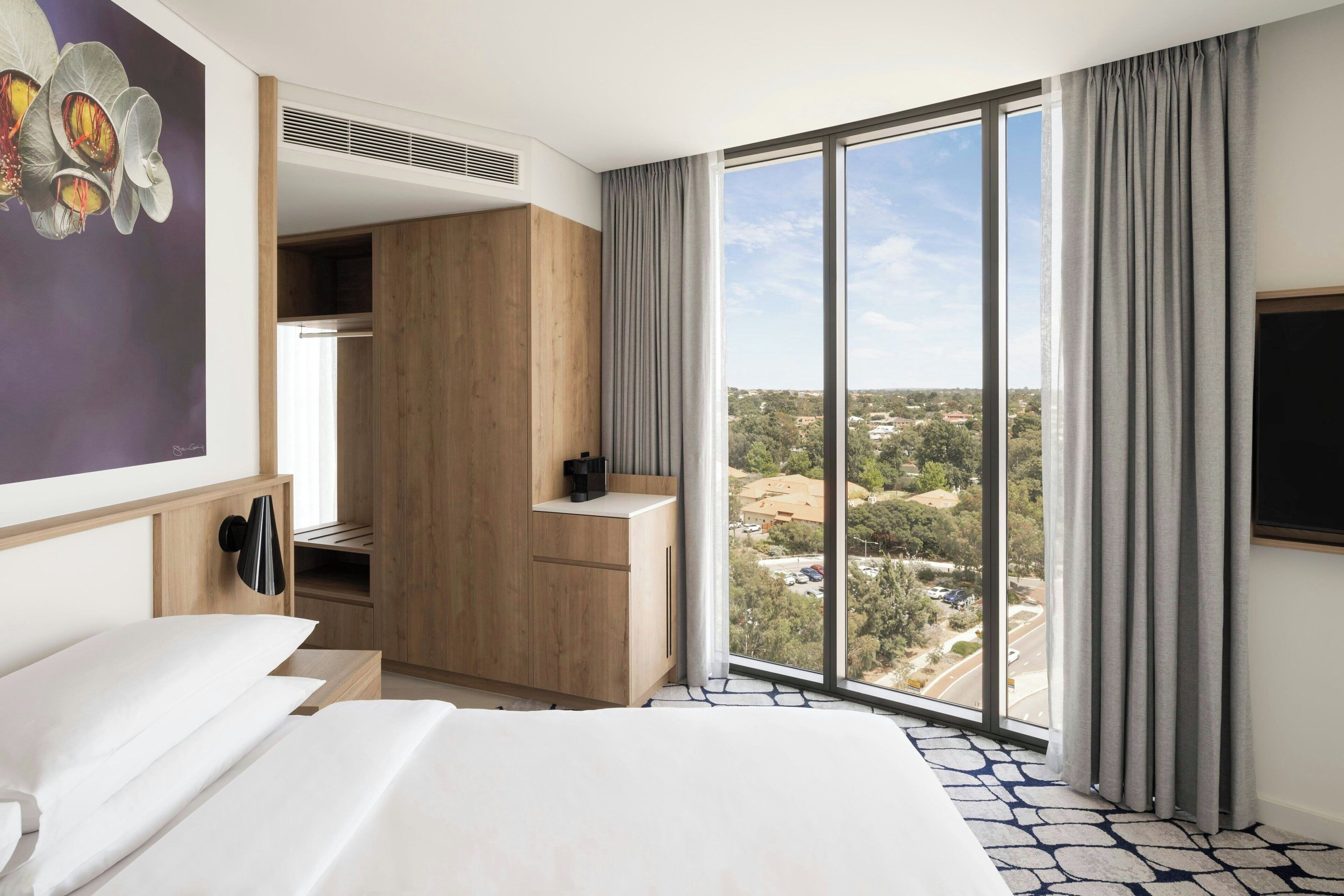 Habitación de hotel con una cama grande, un armario, un escritorio y vistas de la ciudad a través de amplios ventanales.