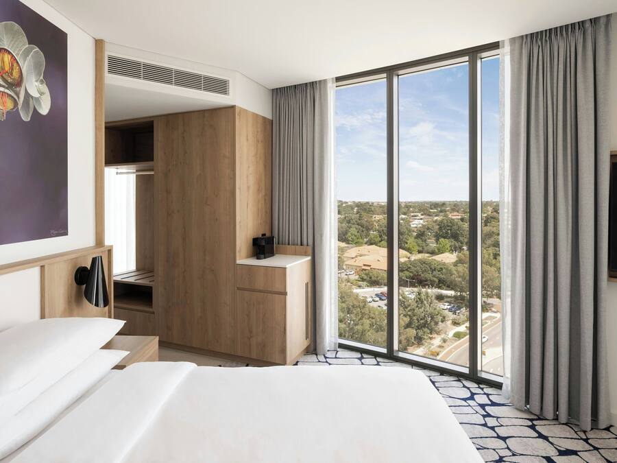 Habitación de hotel con una cama grande, un armario, un escritorio y vistas de la ciudad a través de amplios ventanales.