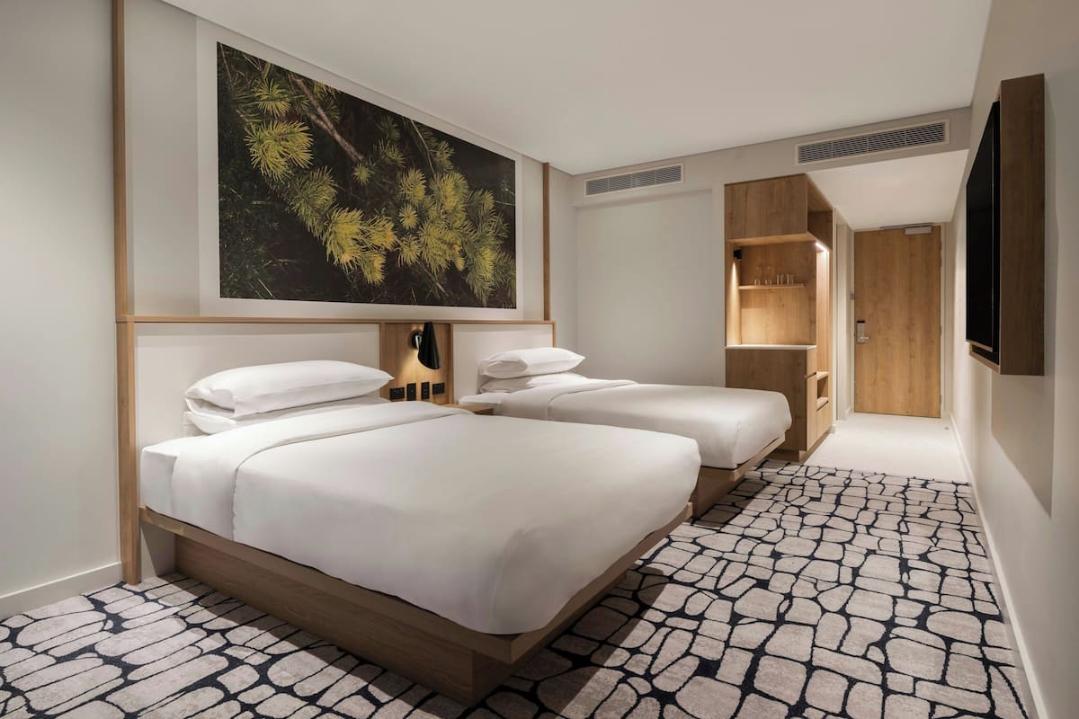 Quarto de hotel com duas camas, um quadro grande com tema de floresta, um guarda-roupa de madeira e uma televisão.