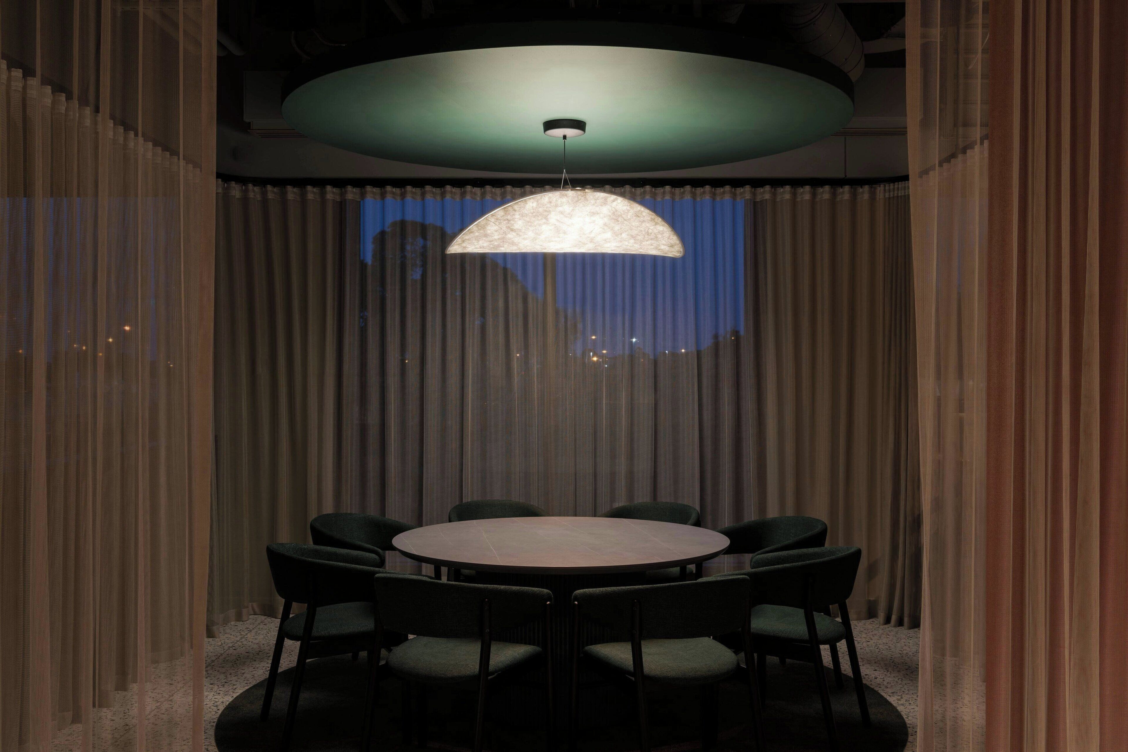 A conference room with a round table and six chairs, a large hanging light fixture, and a view through sheer curtains.