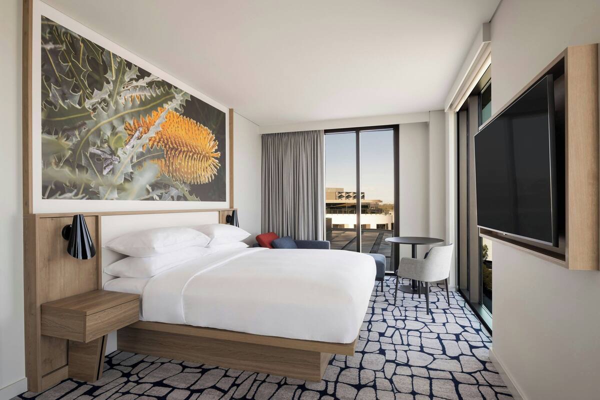 A hotel room with a large bed, a desk, a chair, a TV, and a large window with a city view.