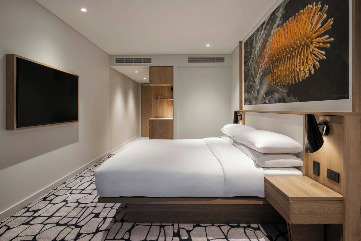 Quarto de hotel moderno com uma cama grande, televisão de tela plana, mesa de cabeceira de madeira, escrivaninha e um quadro de uma flor.