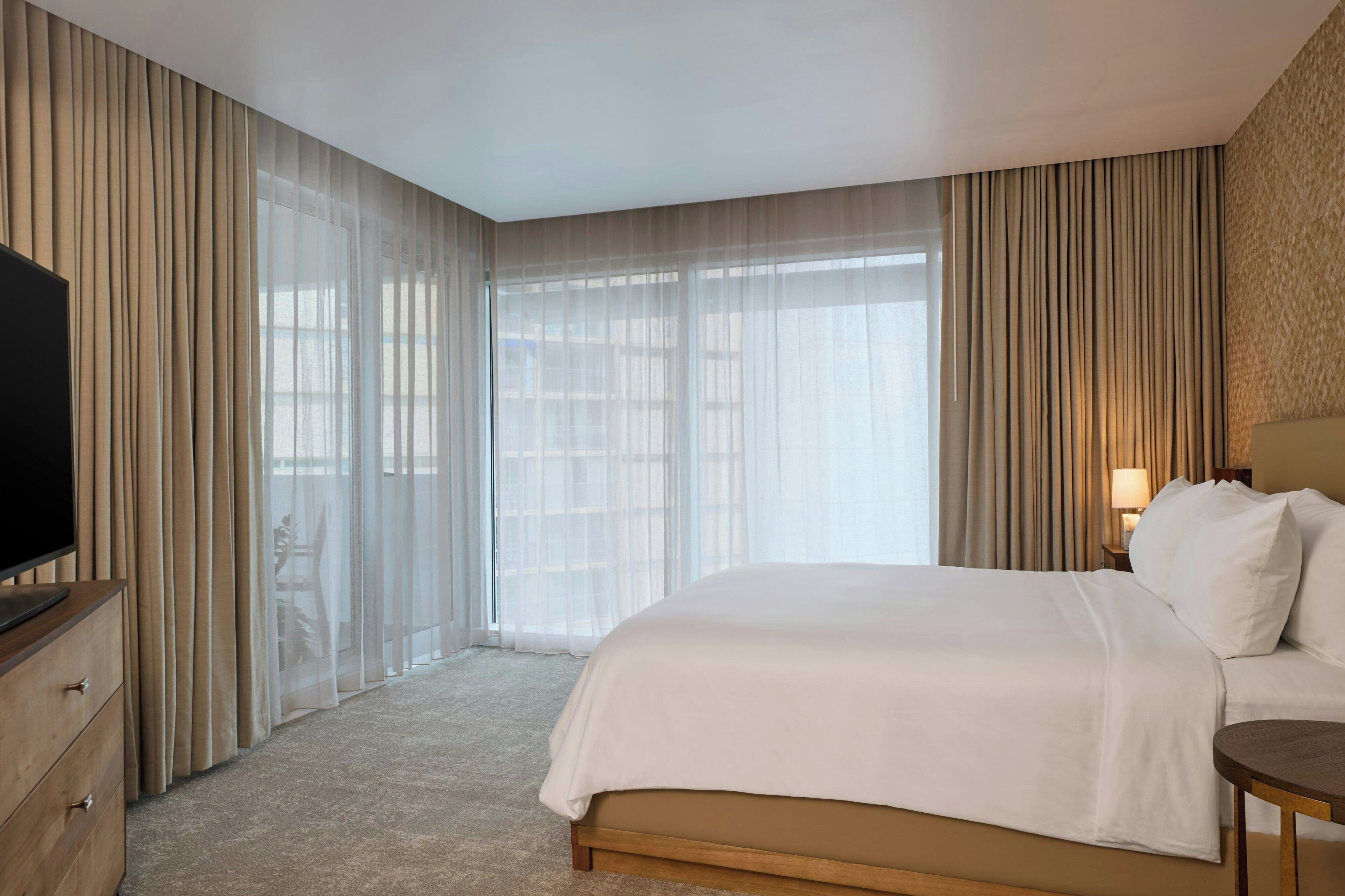 Une chambre d’hôtel avec un grand lit, un téléviseur posé sur une commode en bois et une fenêtre ornée de rideaux légers.