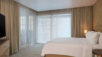 Una habitación de hotel con una cama grande, un televisor sobre un aparador de madera y una ventana con cortinas translúcidas.