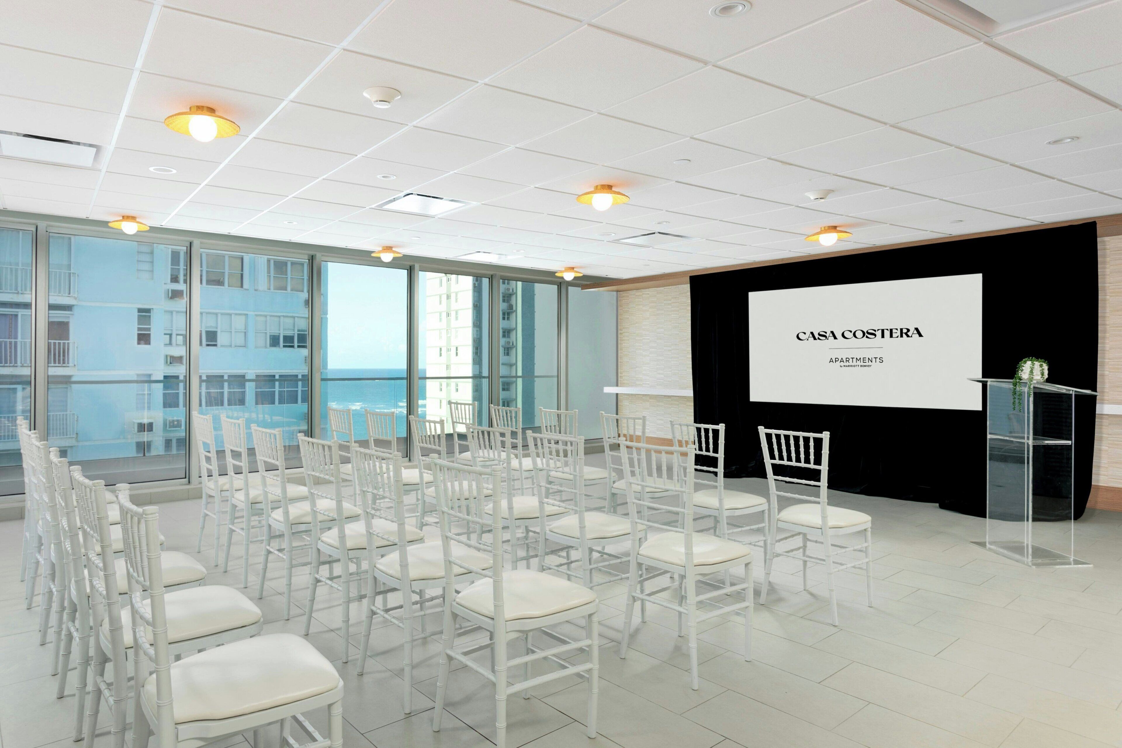 Une salle de conférence avec des chaises blanches disposées en rangées, un pupitre et un écran de projection affichant le texte « CASA COSTERA APARTMENTS ».