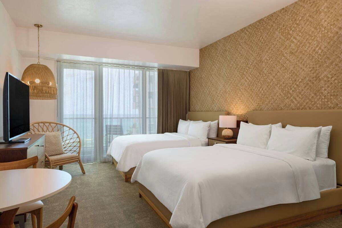A hotel room with two beds, a television, a dining table, and a wicker chair.