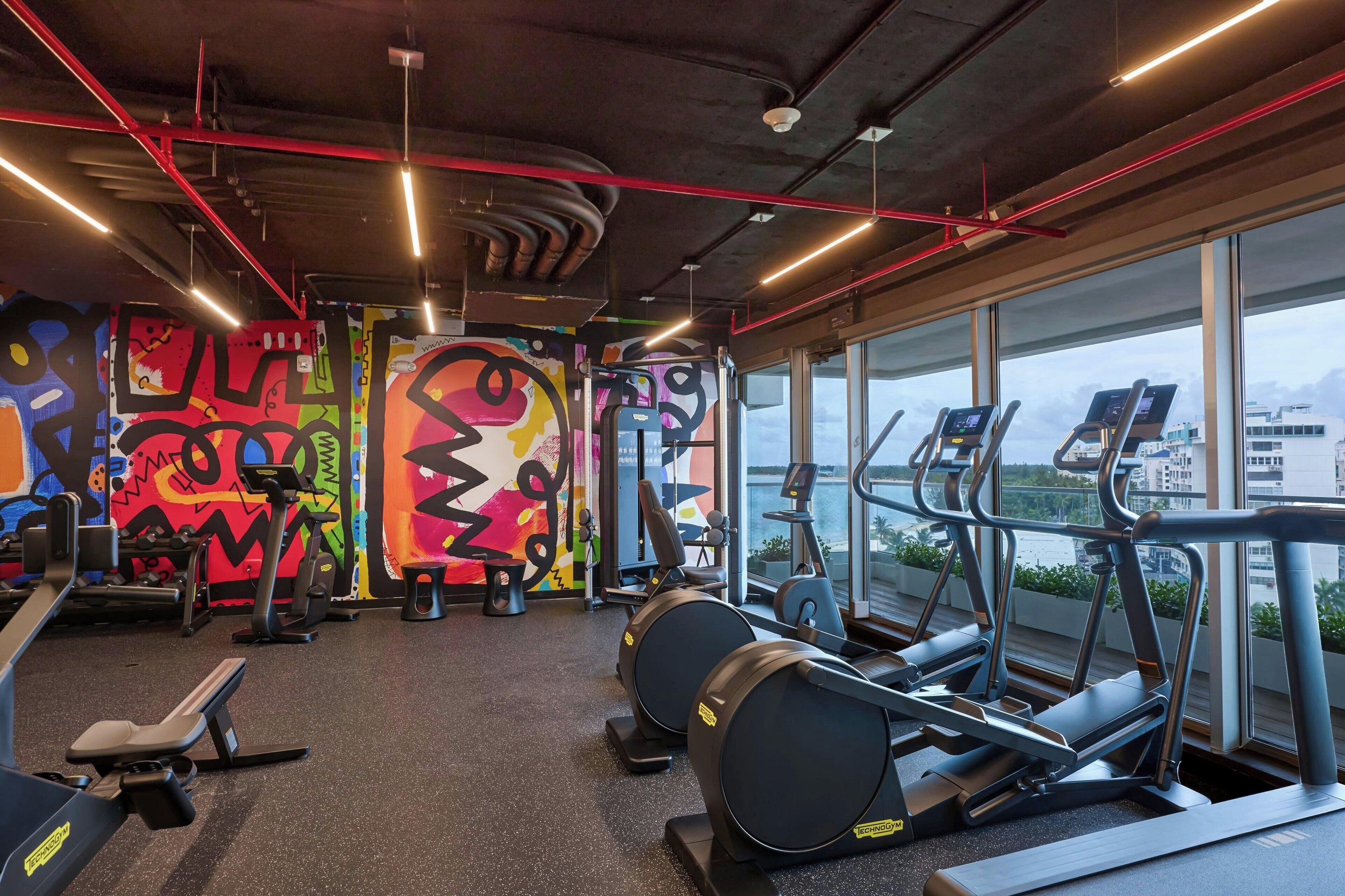 A gym with exercise equipment, a large window with a view, and colorful graffiti on the walls.