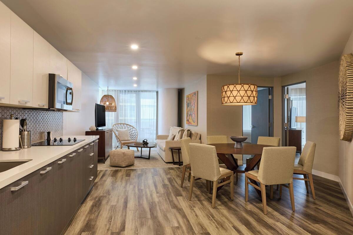 A modern kitchen with a central island, dining area with wooden chairs and a table, and a living room with a sofa and a TV.