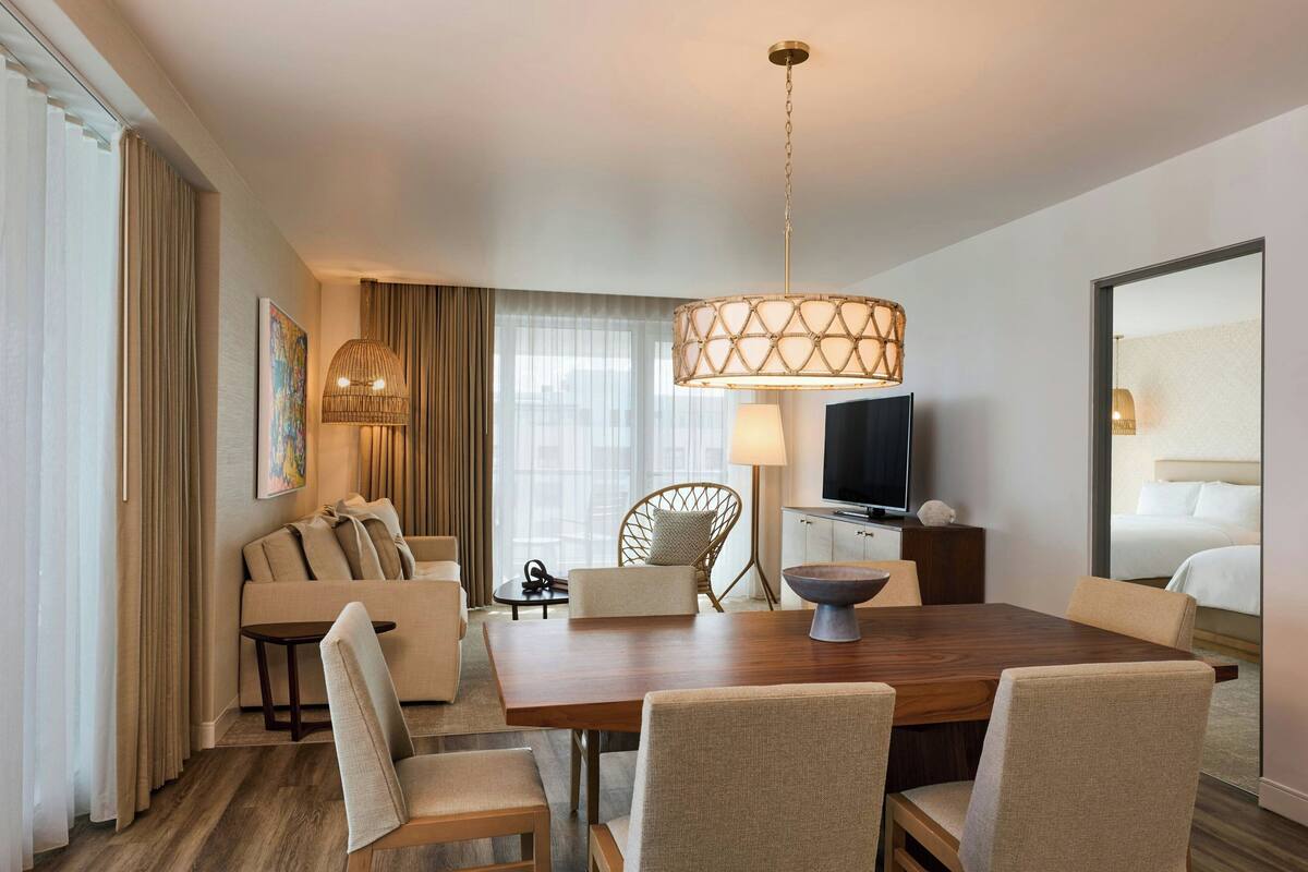 A hotel room with a wooden dining table, beige upholstered chairs, a sofa, a television, and a large mirror.