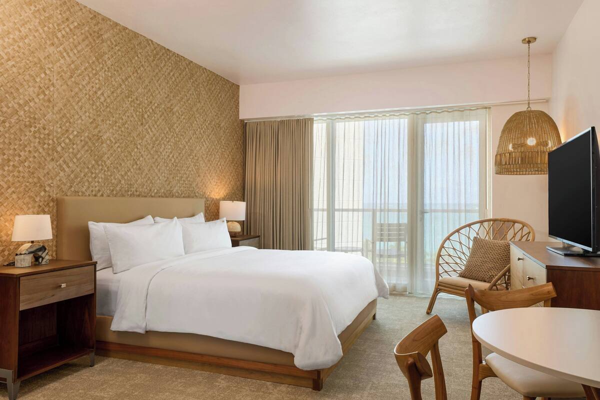 A hotel room with a large bed, a wooden nightstand, a television, a dining table, and a wicker chair.