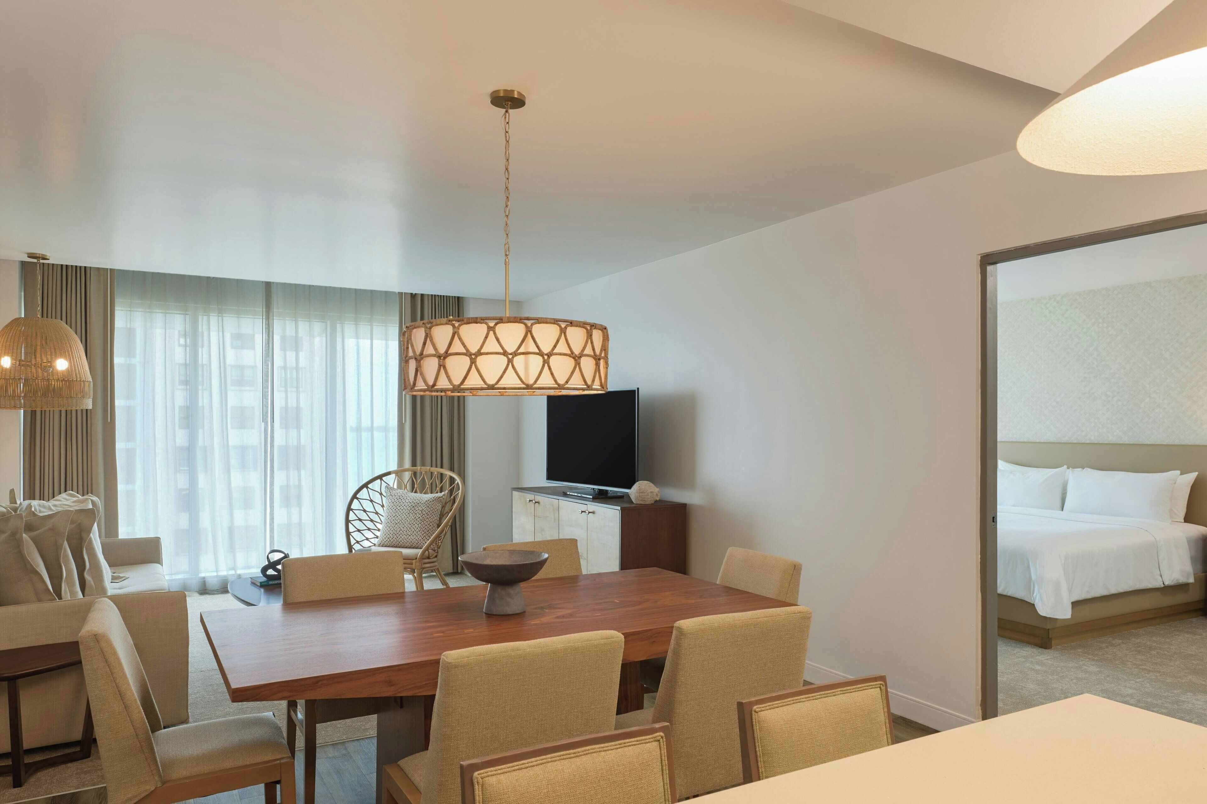 Habitación de hotel con zona de comedor, televisión, cama y un amplio ventanal con cortinas.