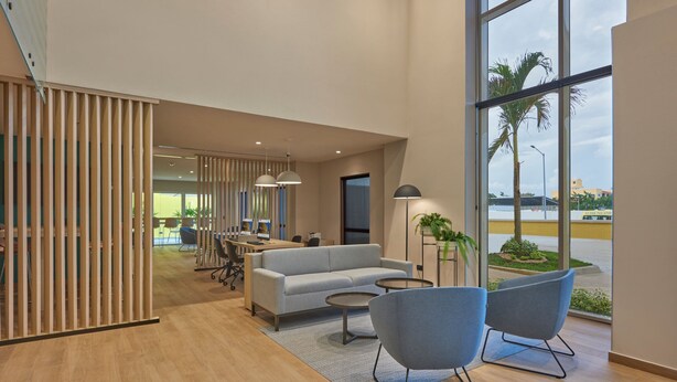 Un moderno vestíbulo de hotel con un sofá, dos sillones, una zona de comedor y grandes ventanales con vistas al exterior.
