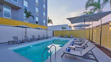 Relax by the pool surrounded by lounge chairs and umbrellas, with a low wall and palm trees in the background.