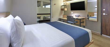 Habitación de hotel con cama, escritorio, silla, televisor y espejo.