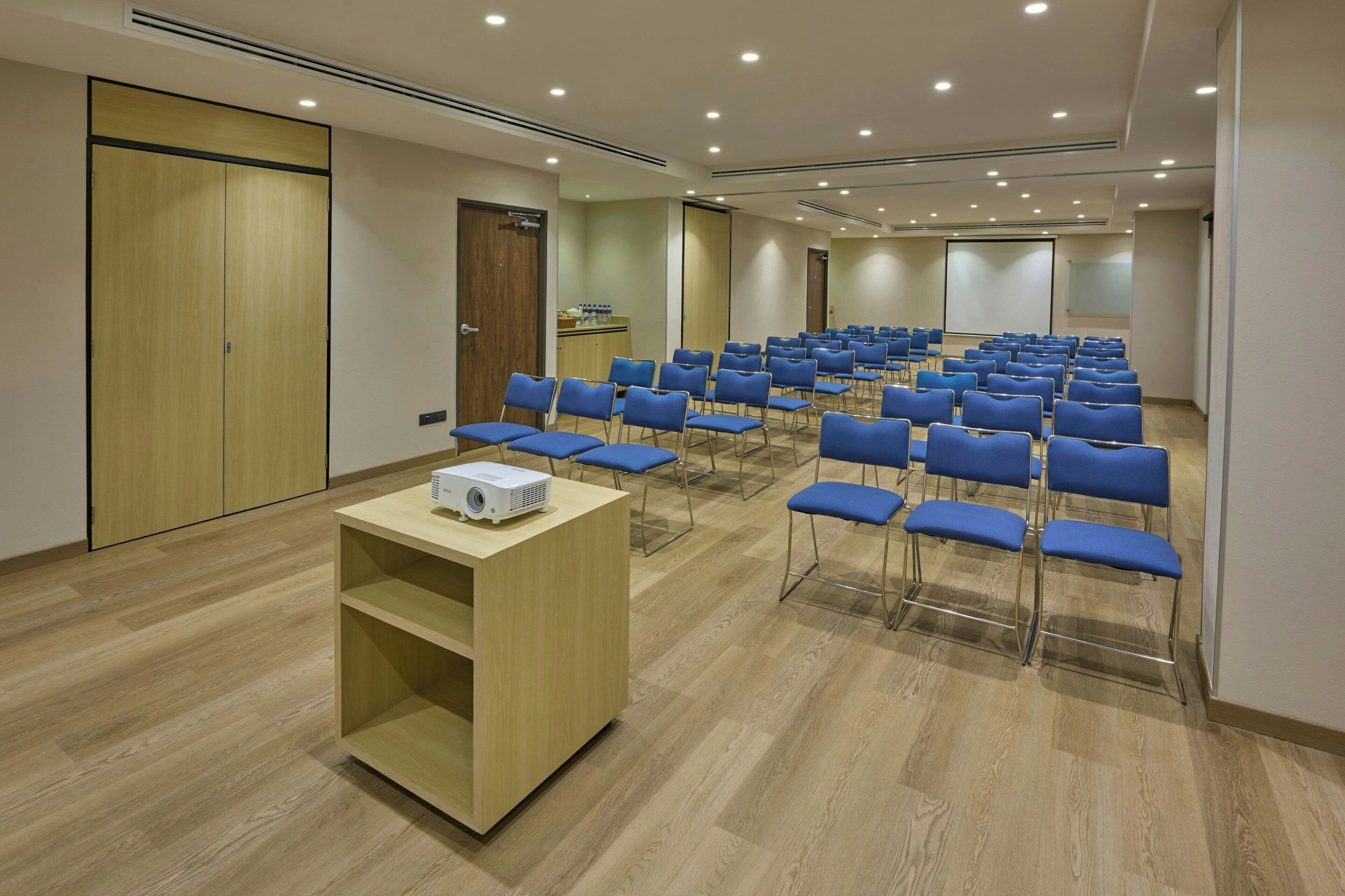 A conference room with rows of blue chairs, a wooden podium, and a projector on a table.