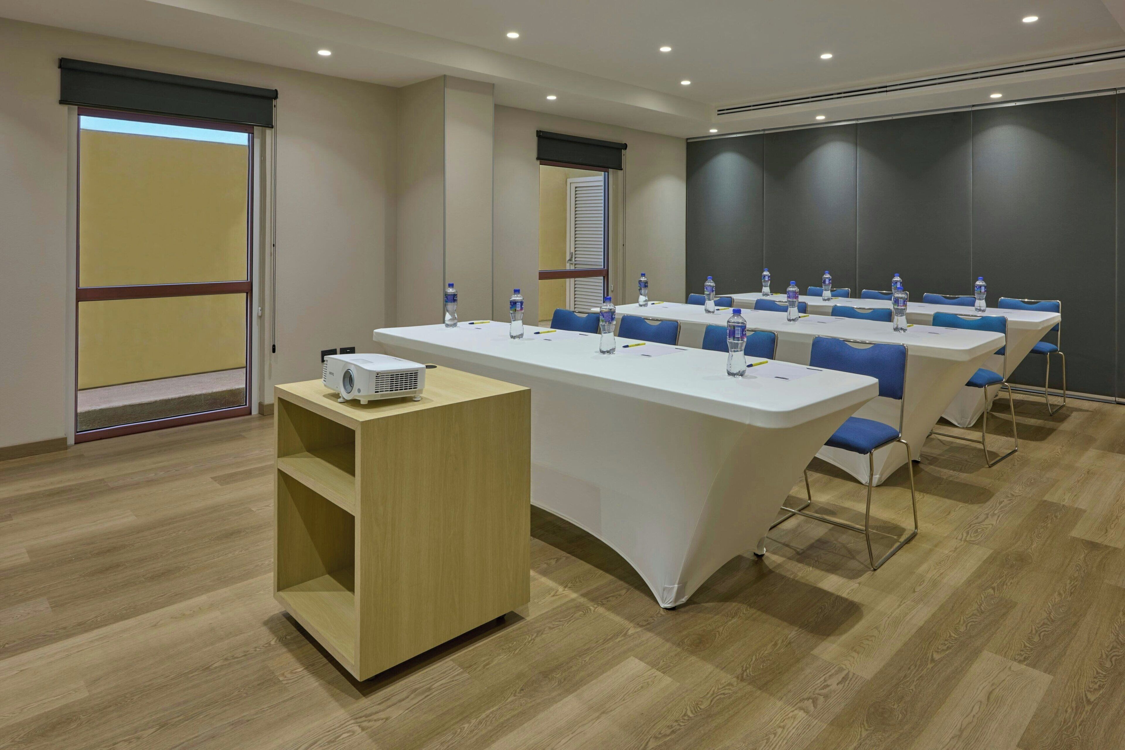 Une salle de conférence avec une longue table, des chaises bleues, des bouteilles d’eau, un projecteur et un buffet en bois.