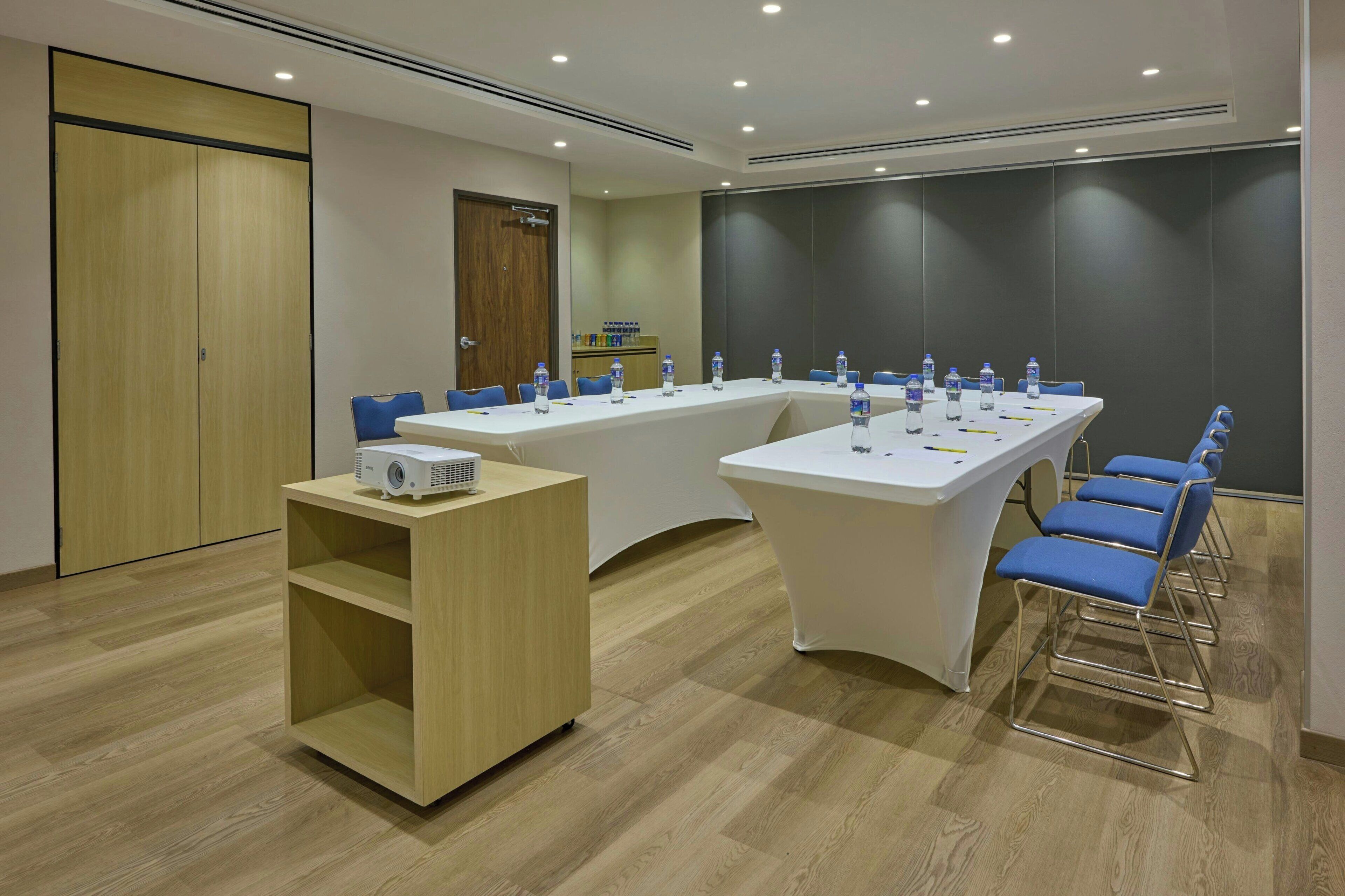 A conference room with a long table, blue chairs, a white tablecloth, and a projector on a stand.