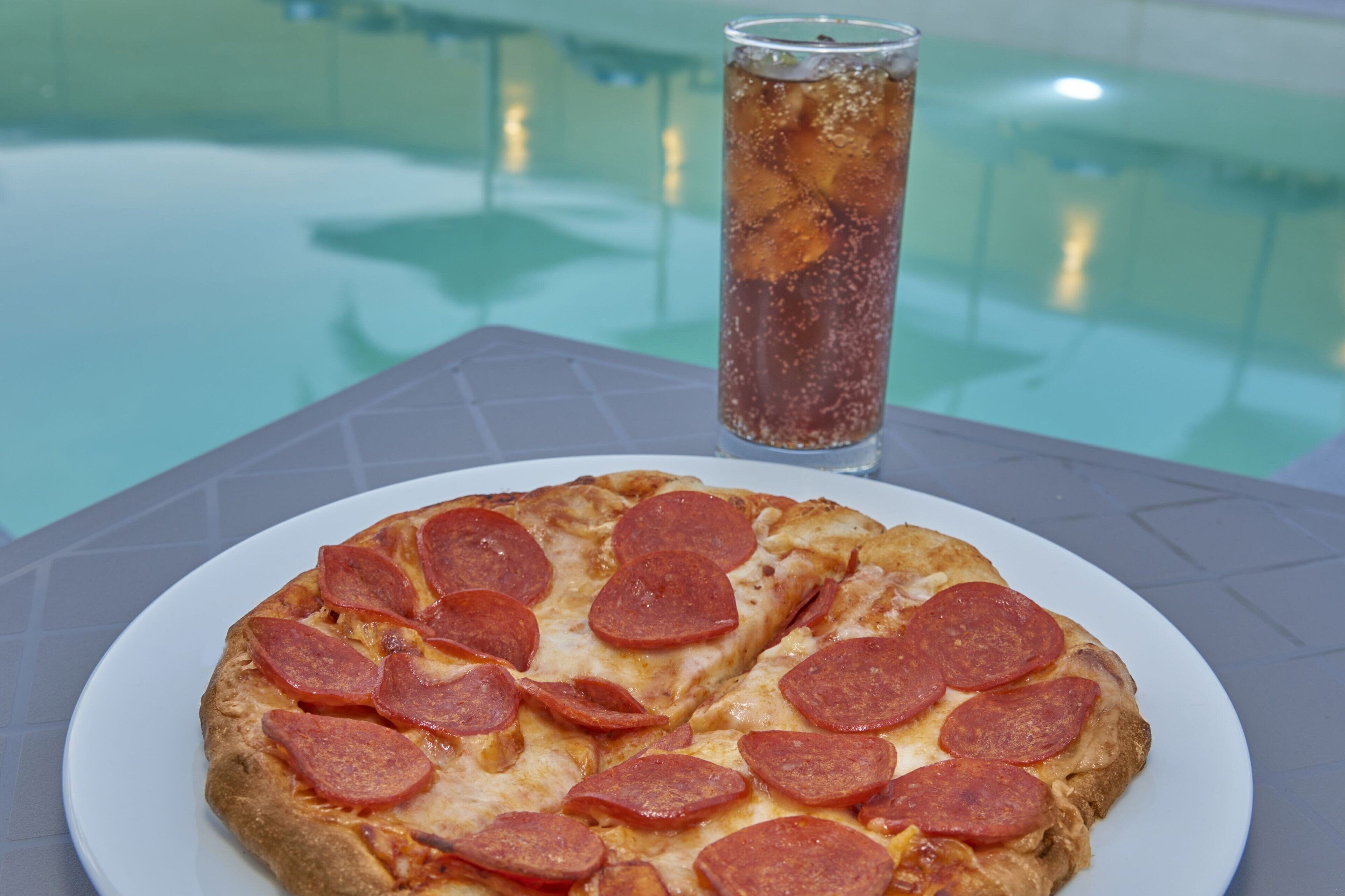 Pizza con rodajas de pepperoni sobre un plato blanco, un vaso de té helado y una piscina al fondo.