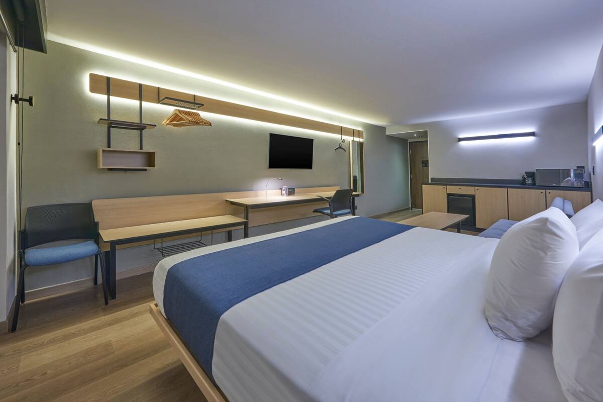 A modern hotel room with a large bed, a desk, a chair, a TV, and a kitchenette.