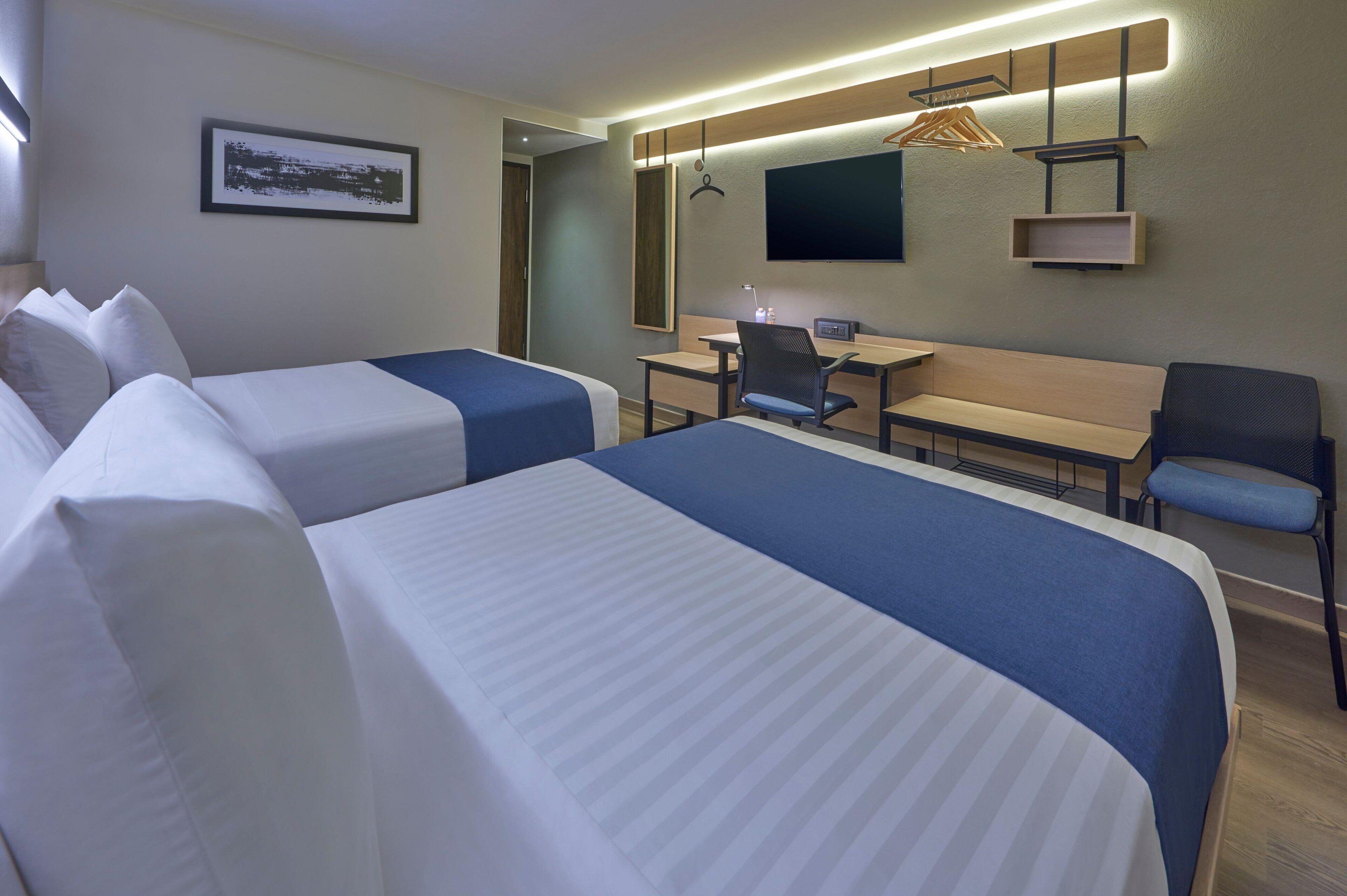 Une chambre d’hôtel avec deux lits, un bureau, une chaise, un téléviseur et un banc en bois.