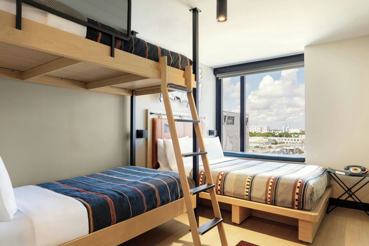 A modern room with a bunk bed, a ladder, a desk, and a large window with a city view.